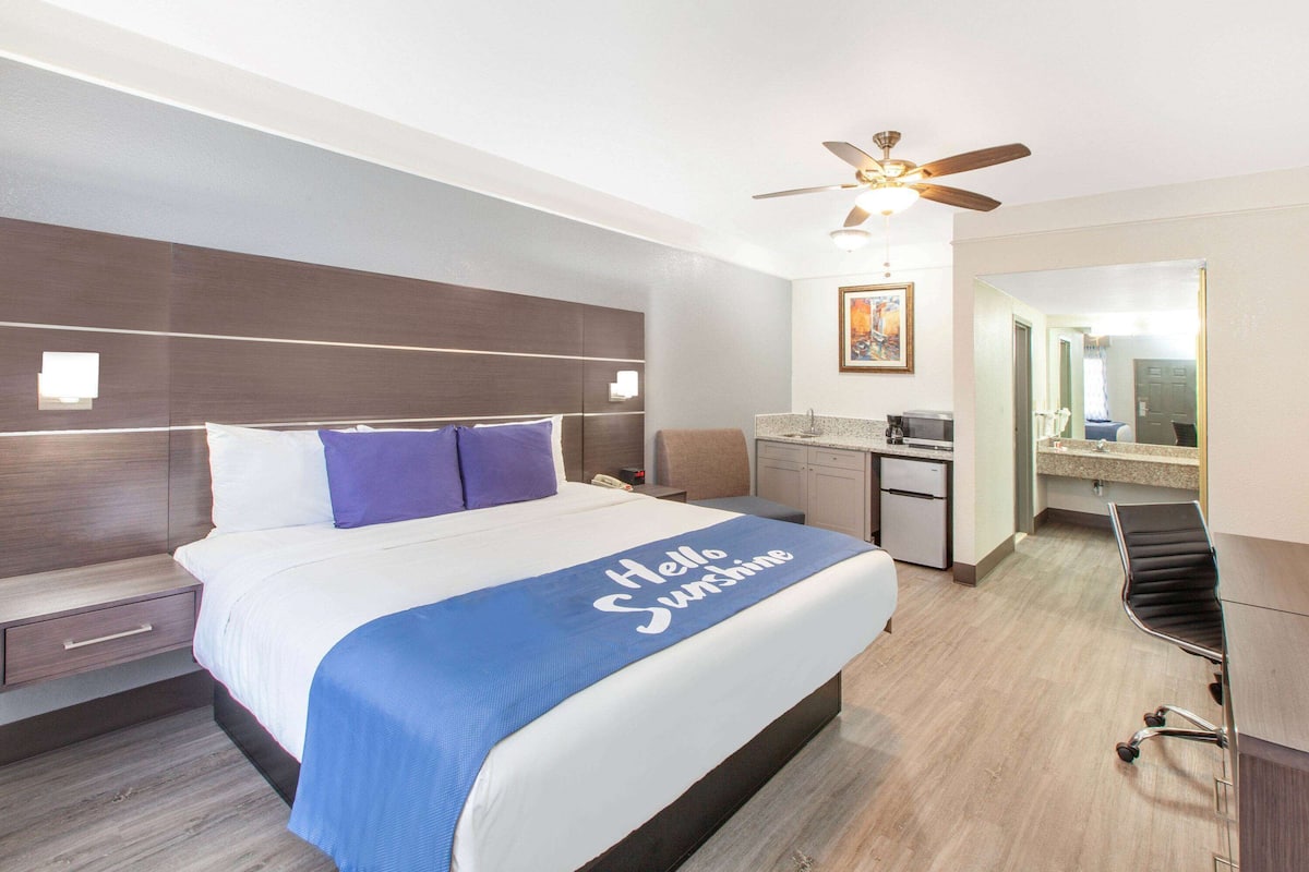 A modern hotel room with a large bed, a desk, a chair, a ceiling fan, and a bathroom with a sink and mirror.
