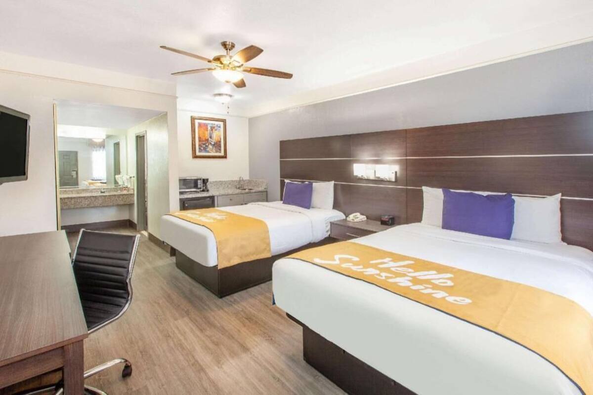 A hotel room with two beds, a desk, a chair, a ceiling fan, a mirror, and a kitchenette.