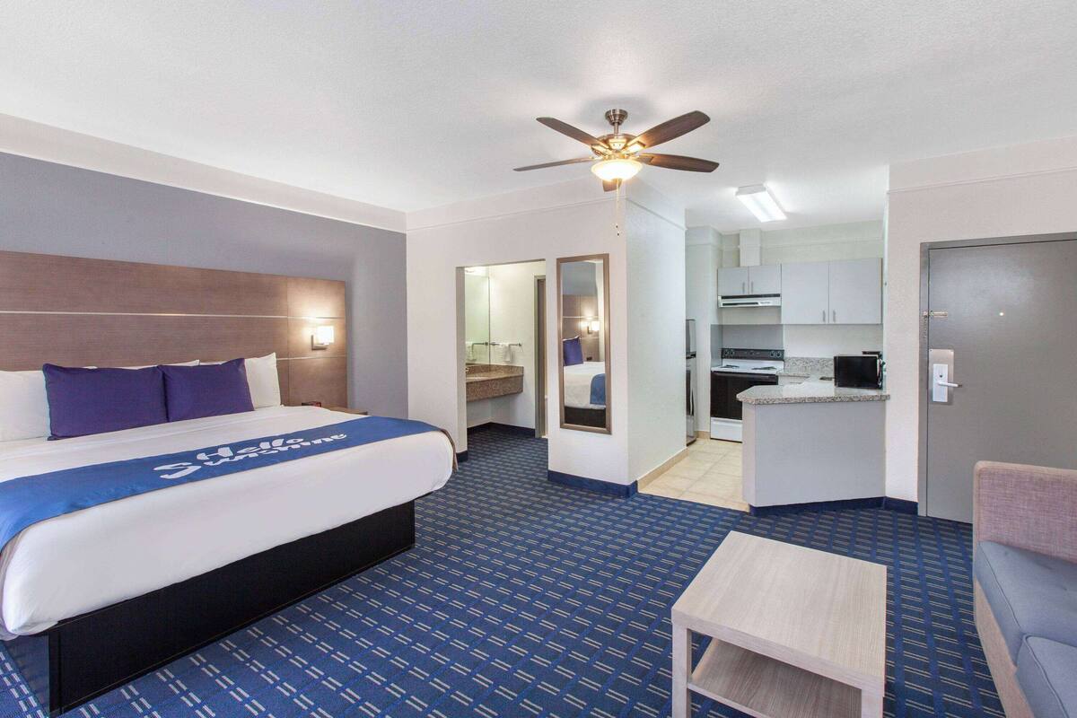 A hotel room with a bed, a desk, a chair, a ceiling fan, and a kitchenette.