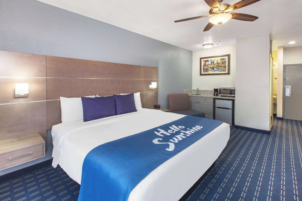A hotel room with a bed, a desk, a chair, a ceiling fan, and a painting on the wall.