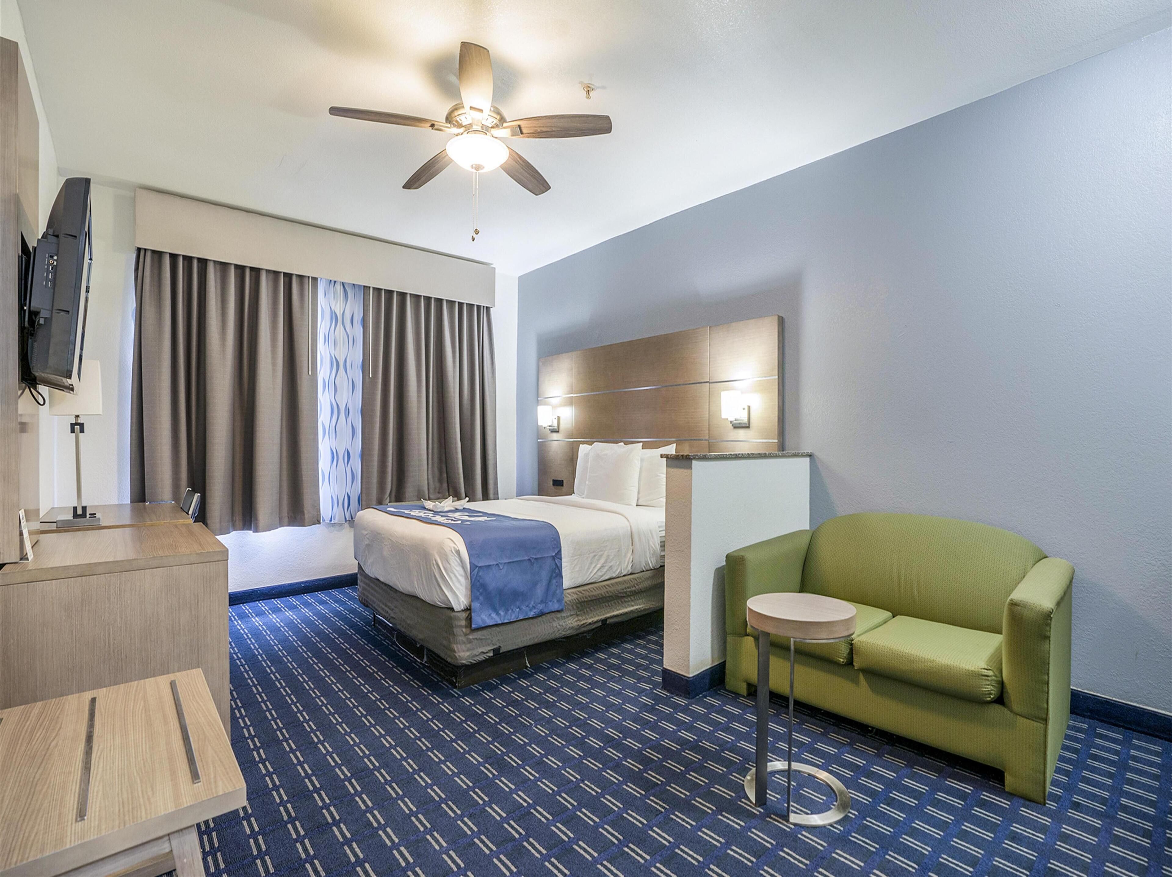 A hotel room with a bed, a desk, a green armchair, a ceiling fan, and a TV.