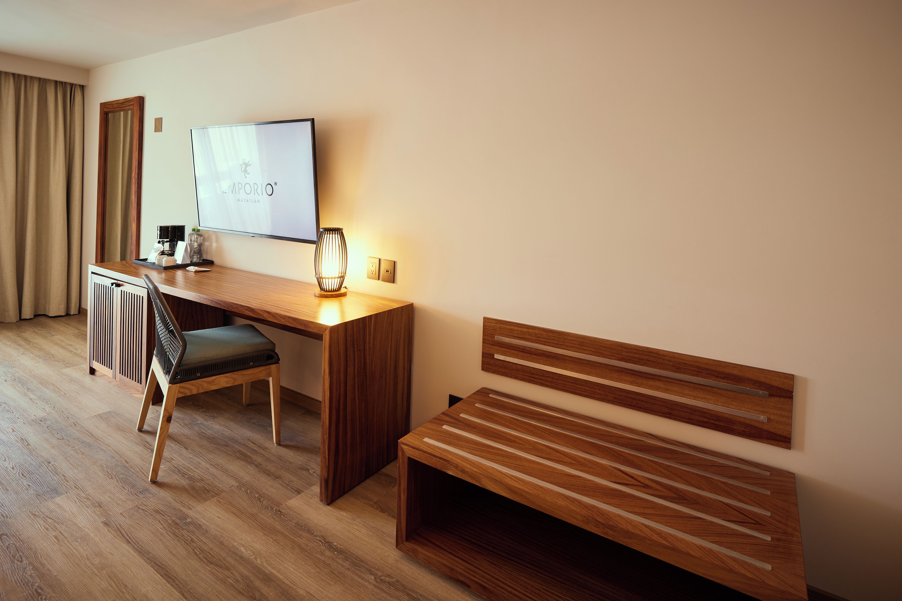 A hotel room with a wooden desk, a chair, a TV, and a bench.