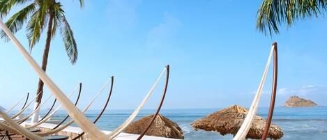 A beach with hammocks and thatched umbrellas, palm trees, and a view of the ocean.