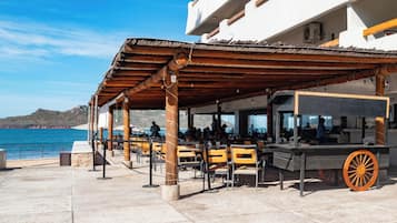 An outdoor seating area with tables and chairs, a covered section with a wooden roof, and a view of the sea and hills in the background.