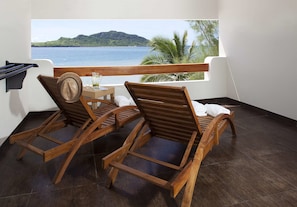 Uma varanda com duas poltronas de madeira, uma mesa pequena com um jarro e vista para um corpo de água e colinas.