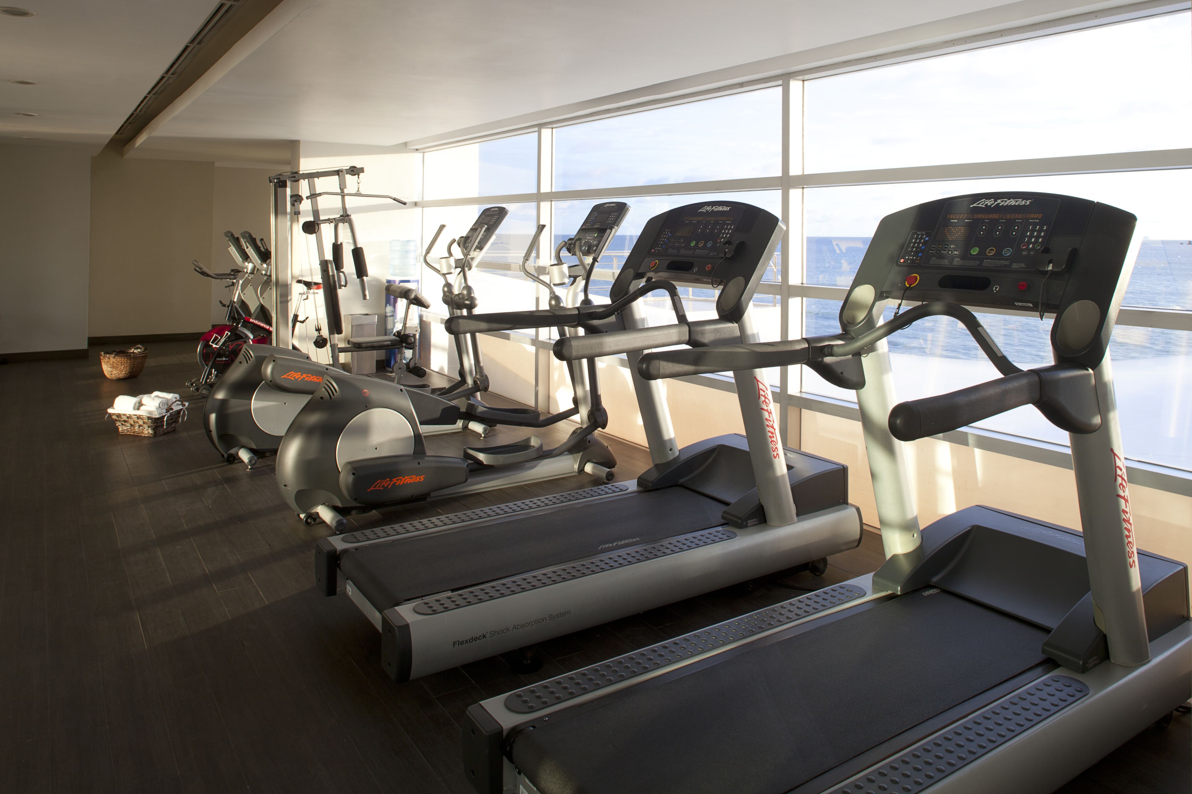 A gym with treadmills, ellipticals, and a weight machine, large windows with a view of the sea, and a basket with towels on the floor.