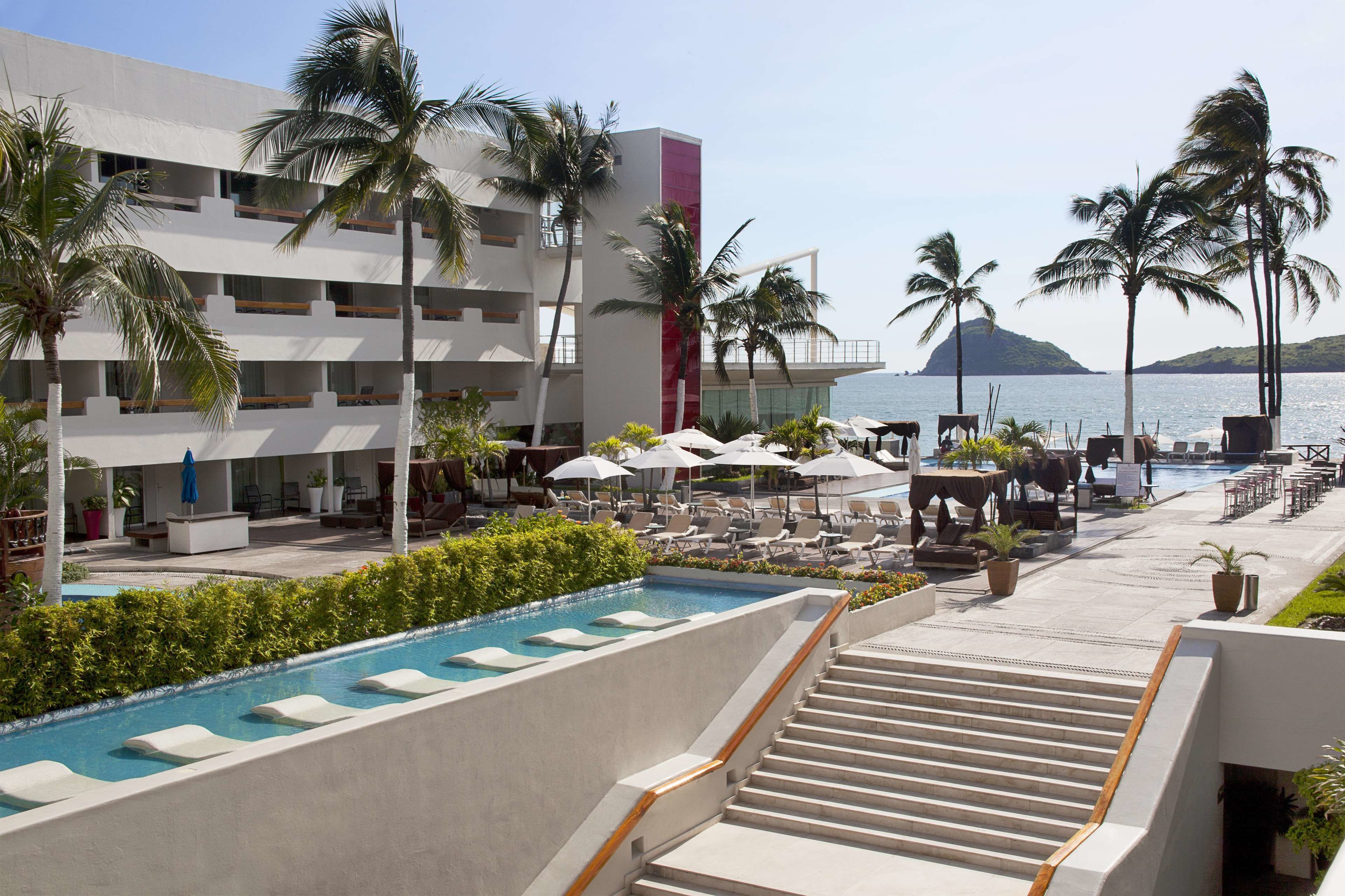 A hotel pool area with lounge chairs, umbrellas, and a staircase leading to a building with balconies overlooking the sea.