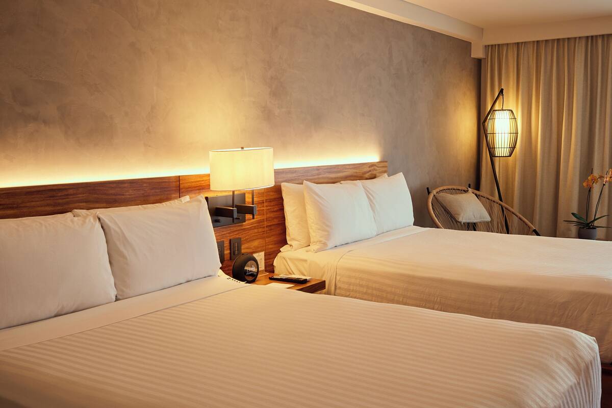 A hotel room with two beds, a nightstand with a lamp, a chair, and a potted plant.