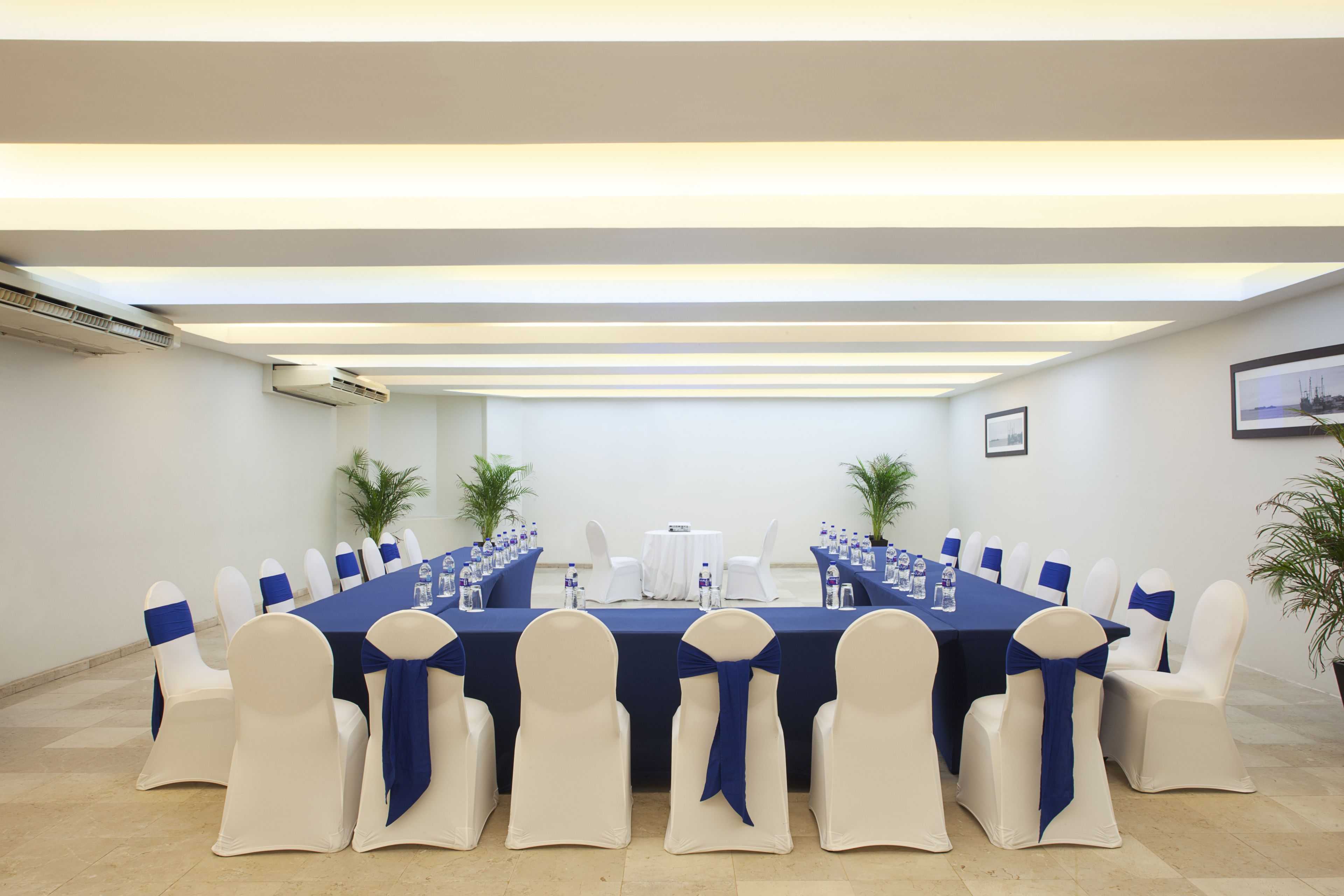 A conference room with a long table, white chairs with blue sashes, and potted plants.