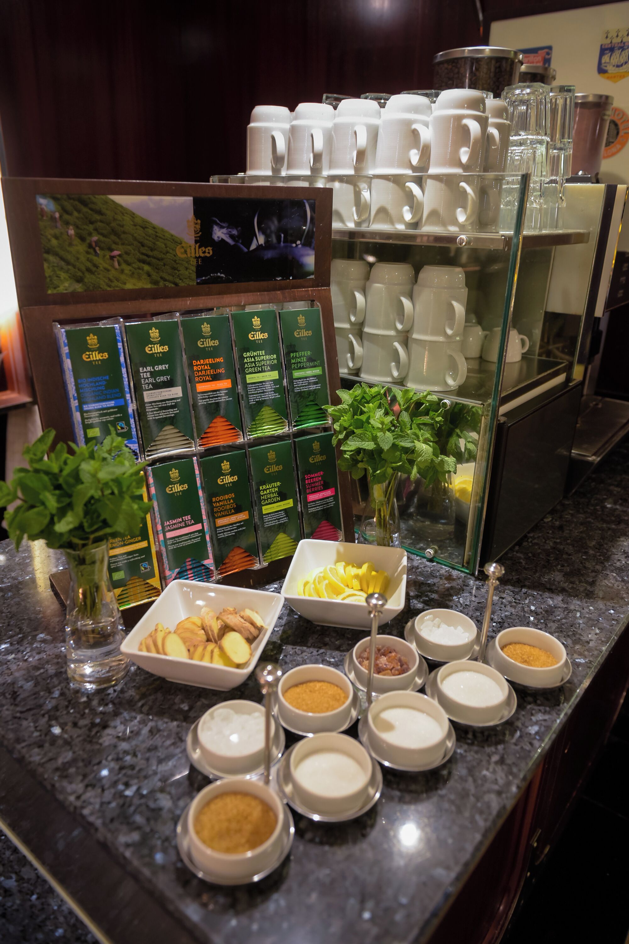 Een koffiehoek met diverse thee- en koffiemogelijkheden, een presentatie van theedozen en een glazen pot met verse muntblaadjes.