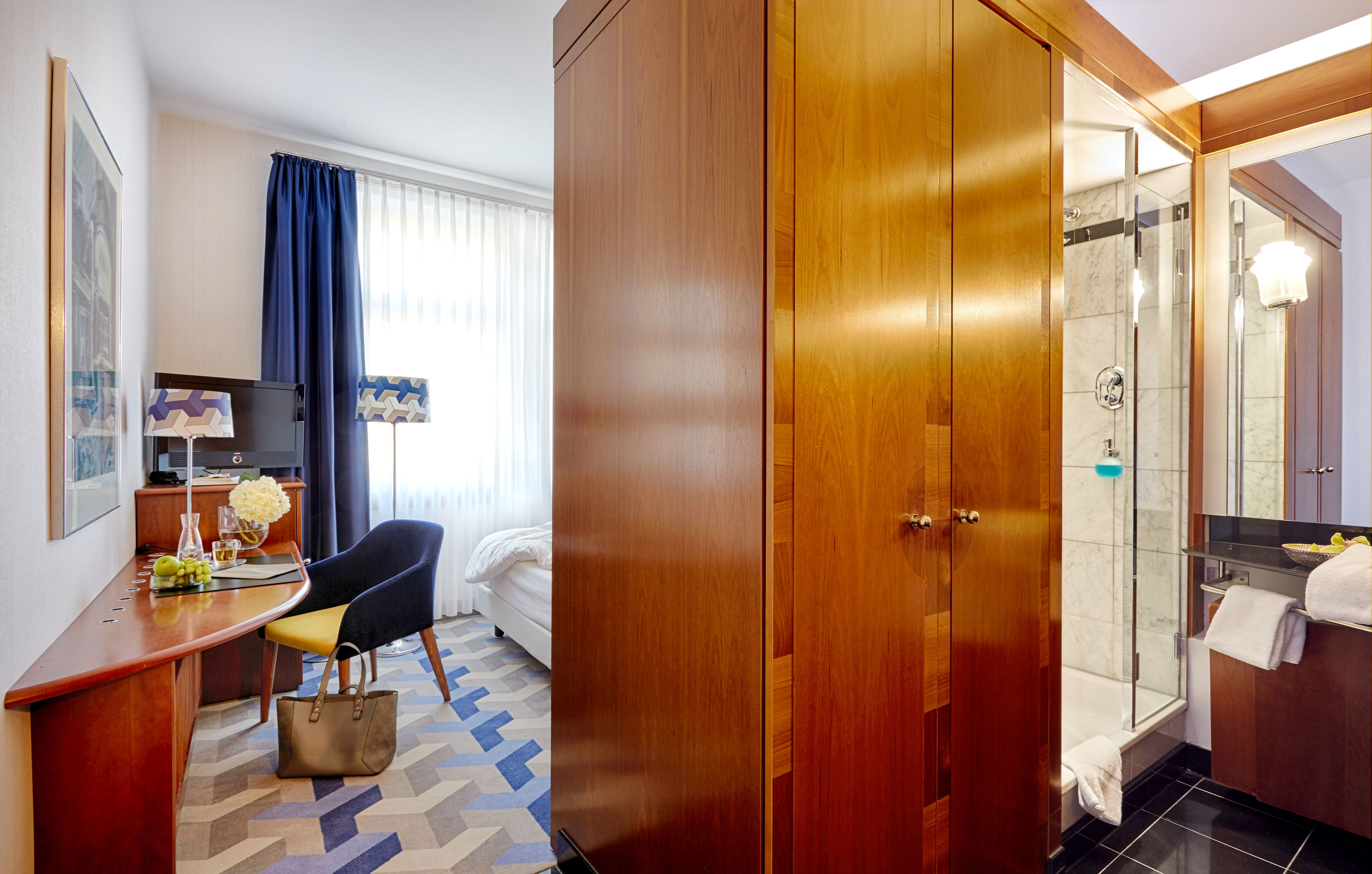 A hotel room with a wooden wardrobe, a desk with a chair, a bed, a shower area, and a bathroom with a mirror and sink.