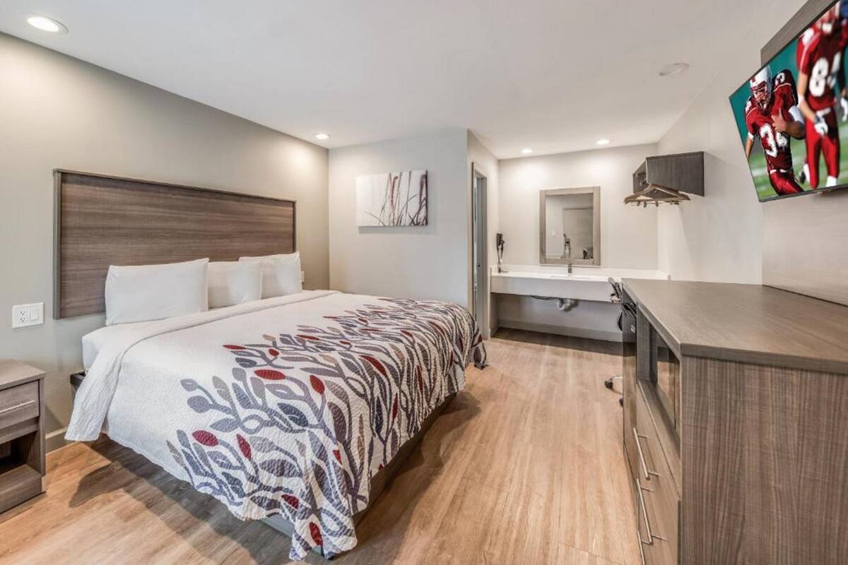A modern hotel room with a large bed, a bedside table, a built-in bench, a large mirror, and a flat-screen TV mounted on the wall.