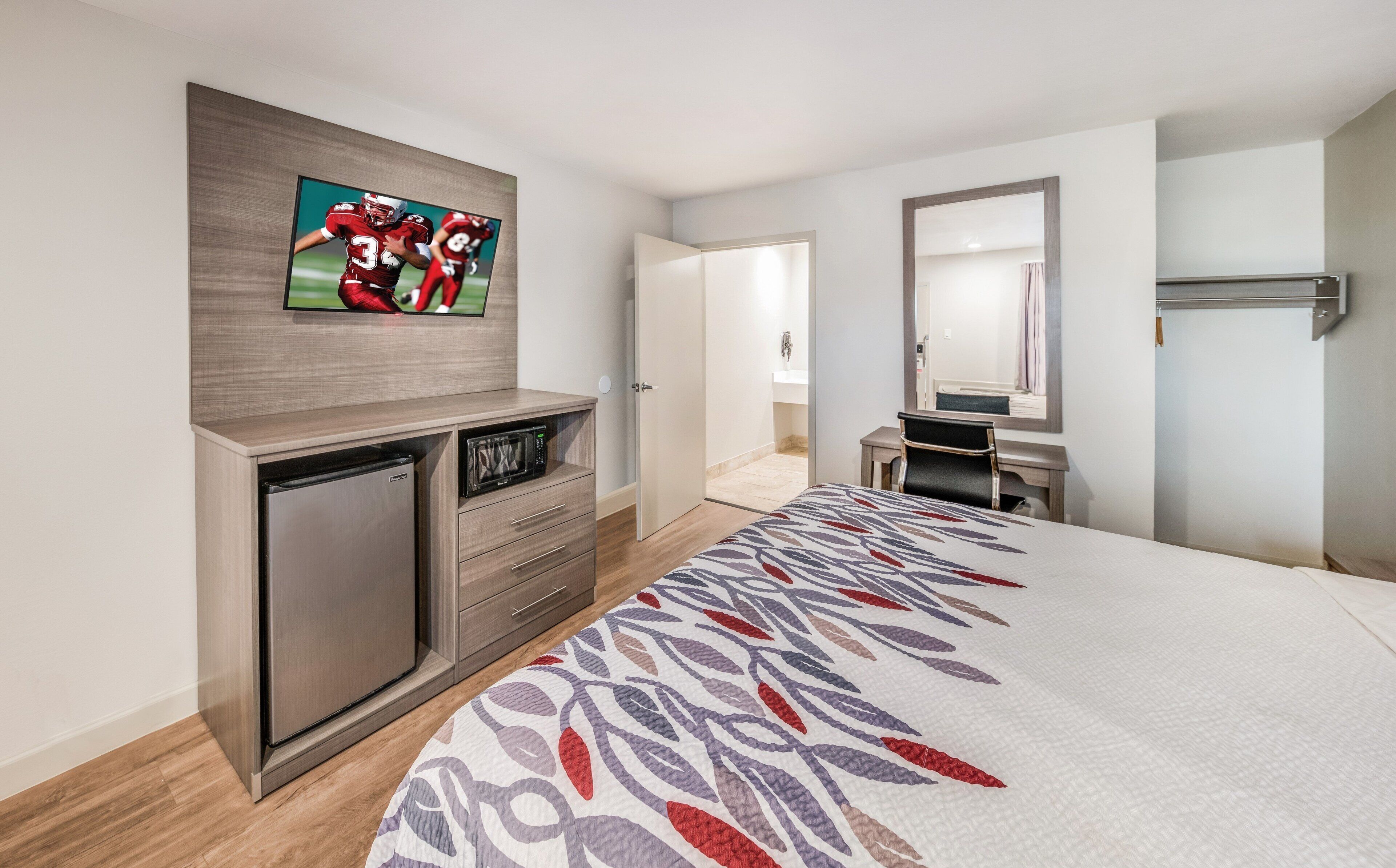 Habitación de hotel con cama, televisor montado en la pared, escritorio con silla, microondas y puerta que da a un baño.