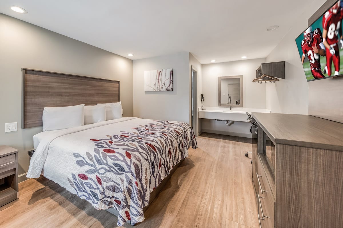 A modern hotel room with a large bed, a bedside table, a built-in TV, a bathroom with a mirror and sink, and a wooden floor.