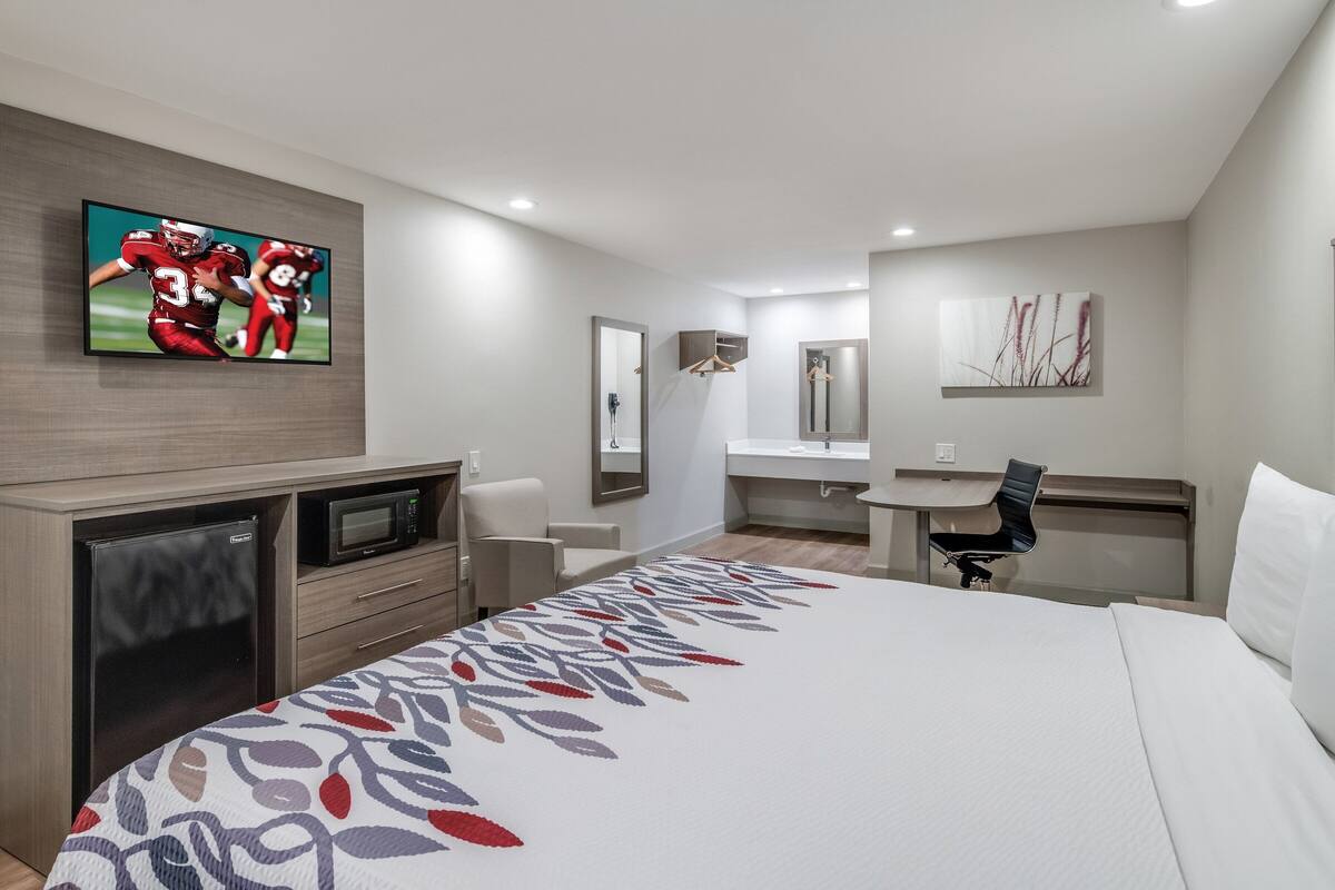 A hotel room with a bed, a desk, a chair, a TV, a microwave, and a bathroom mirror.