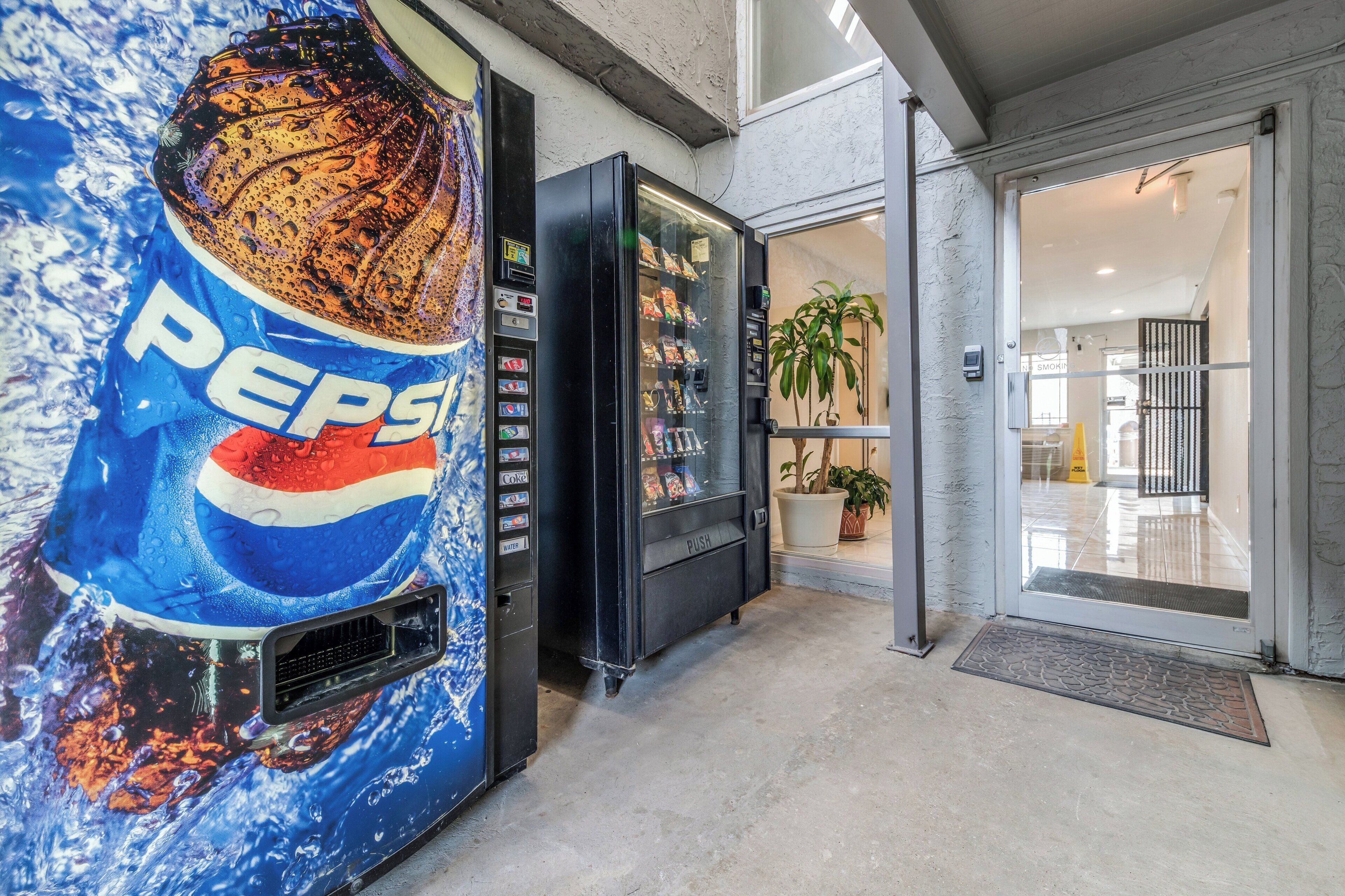 Máquina expendedora con el logotipo de Pepsi, entrada con puerta de vidrio y una planta en maceta en su interior.