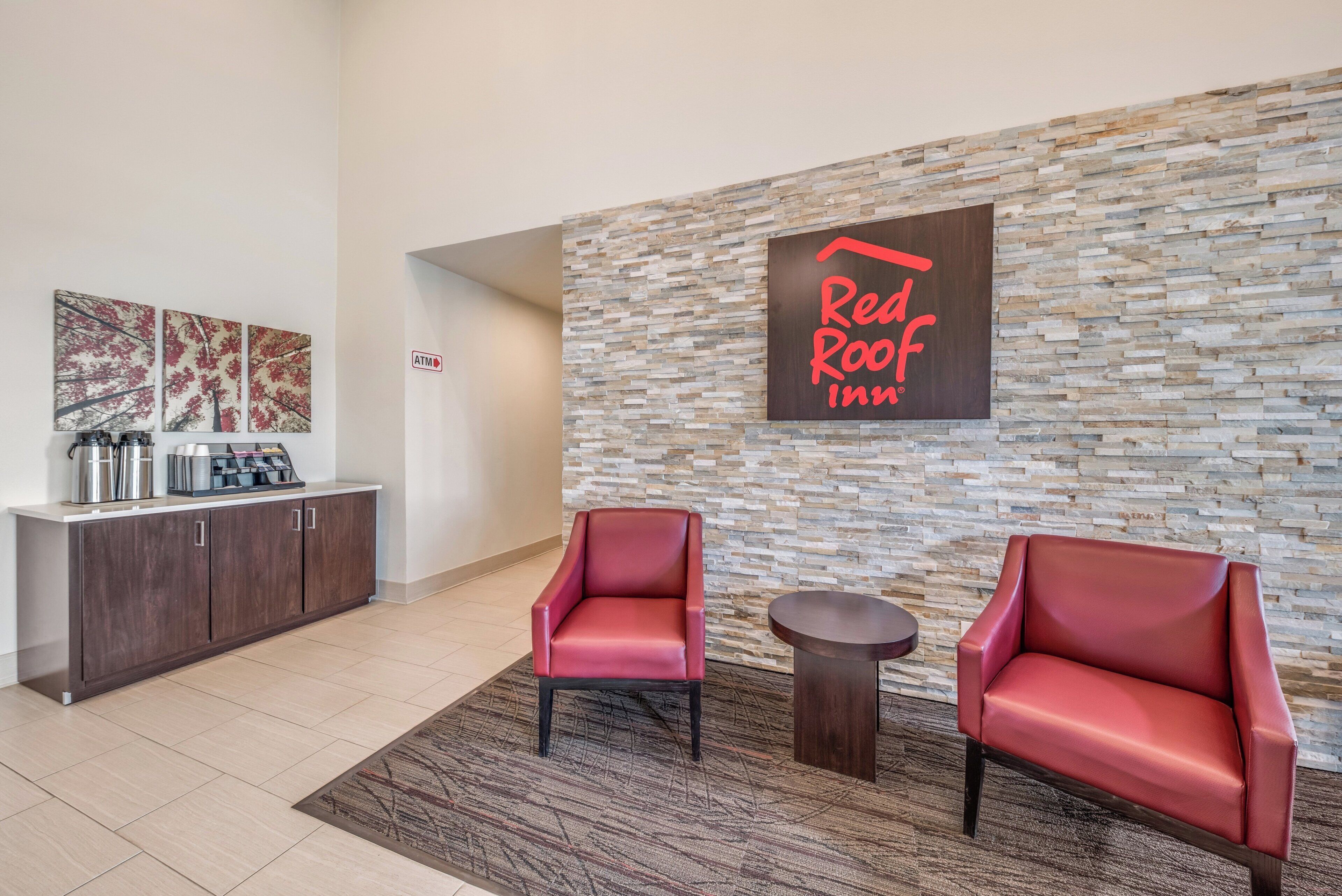 Vestíbulo de Red Roof Inn con pared de piedra, dos sillones de cuero, una pequeña mesa y un mostrador de recepción con máquinas de café.