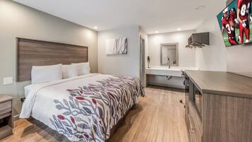 Una habitación de hotel moderna con una cama grande, una mesita de noche, un armario empotrado, un baño con lavamanos y espejo, y un televisor de pantalla plana montado en la pared.