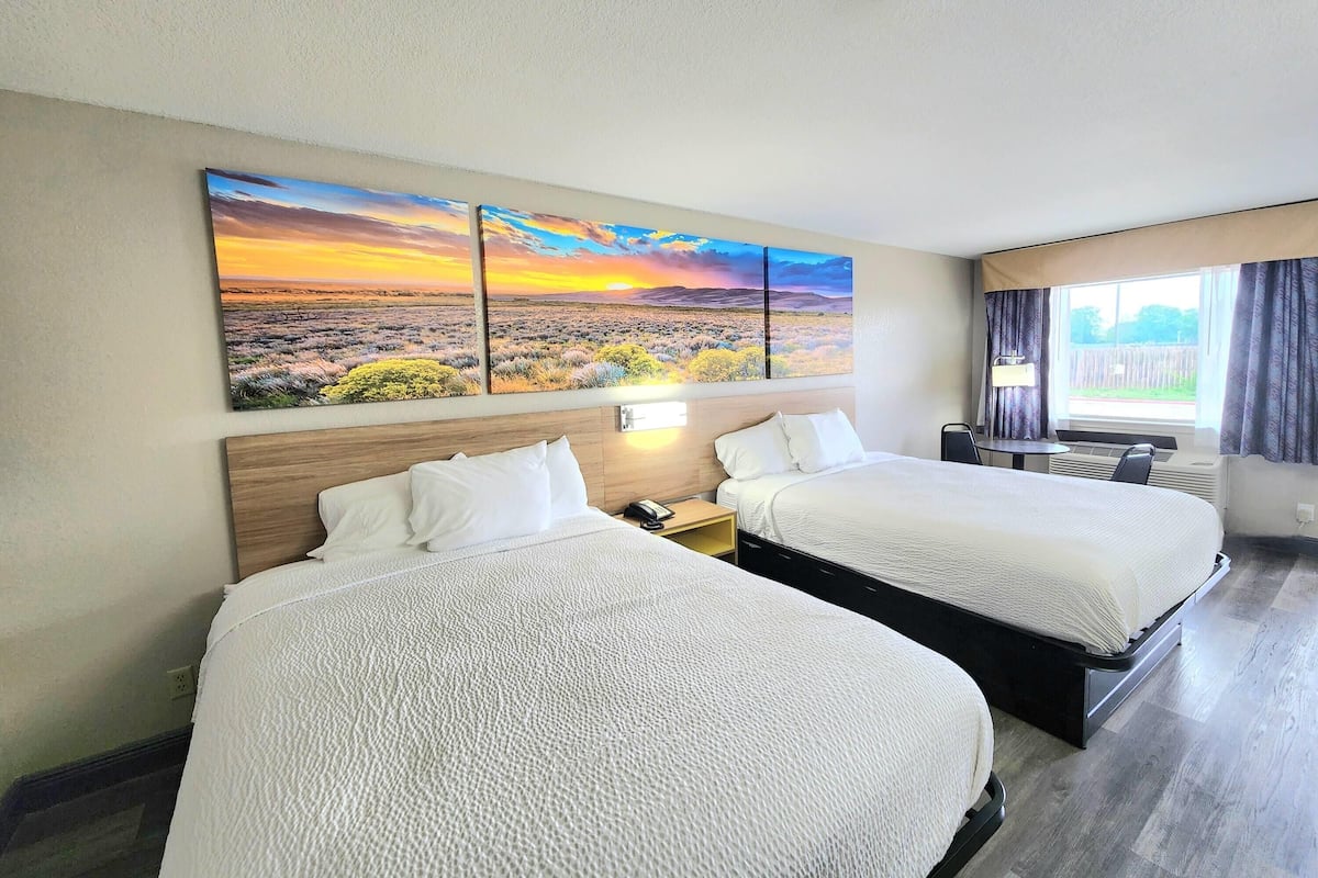 A hotel room with two beds, a painting of a sunset landscape, a desk with a lamp, and a window with curtains.