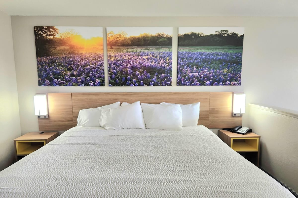 A hotel room with a large bed, white bedding, a wooden headboard, and three wall-mounted paintings of a field at sunset.