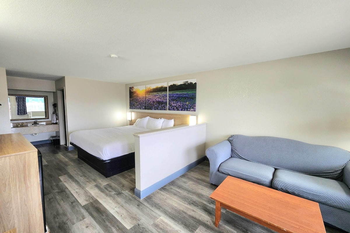 A hotel room with a bed, a sofa, a wooden table, a painting on the wall, and a kitchenette area.