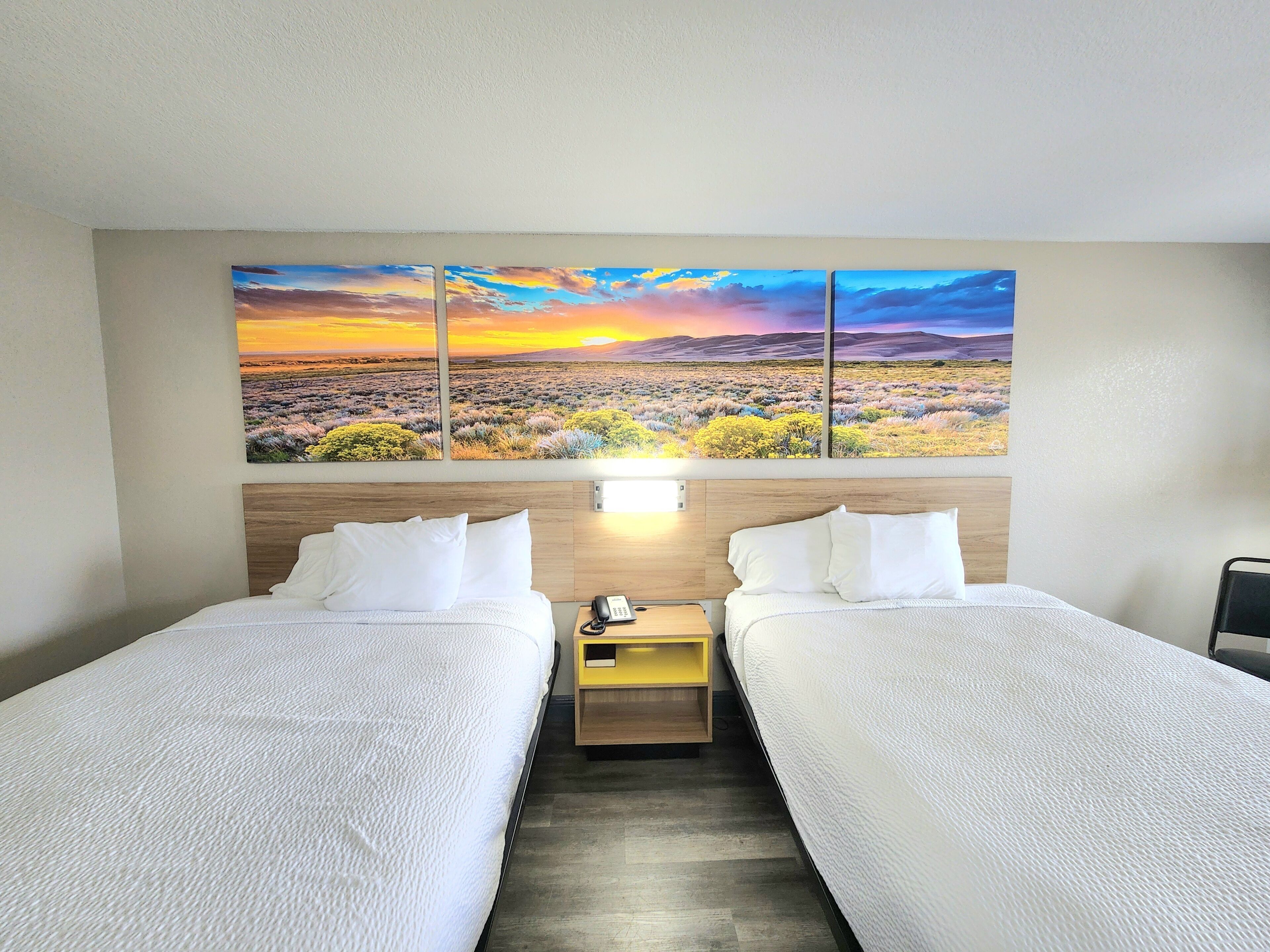 Habitación de hotel con dos camas, una mesita de noche, un teléfono y un cuadro grande de paisaje sobre las camas.