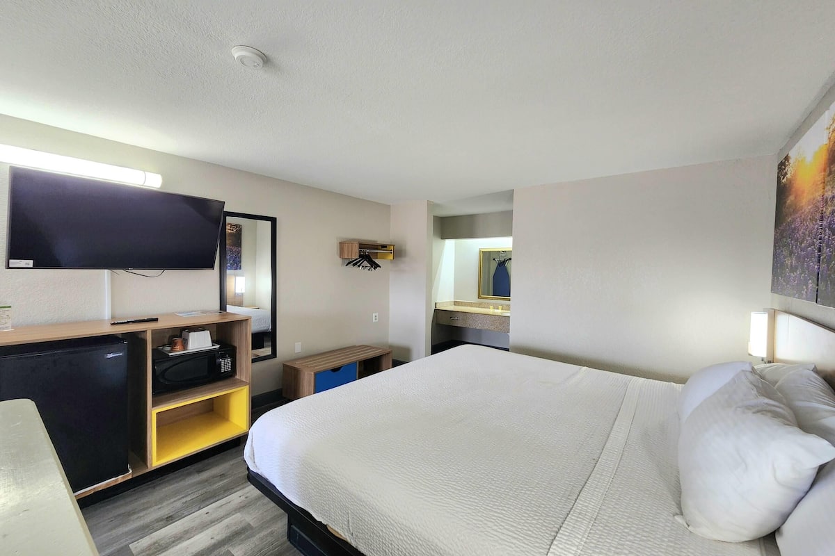 A hotel room with a bed, a flat-screen TV, a microwave, a small refrigerator, a wooden desk, and a mirror.