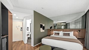 A modern hotel room with a large bed, a wooden headboard, a small bedside table, and a nightstand. There is a bathroom visible through an open door. - Tower Mill Hotel (Spring Hill)