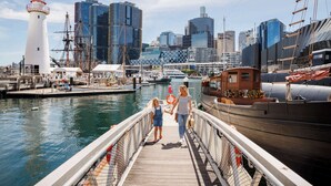 Eine hölzerne Mole, die zu einem Hafen führt, mit einem Leuchtturm und modernen Gebäuden im Hintergrund.