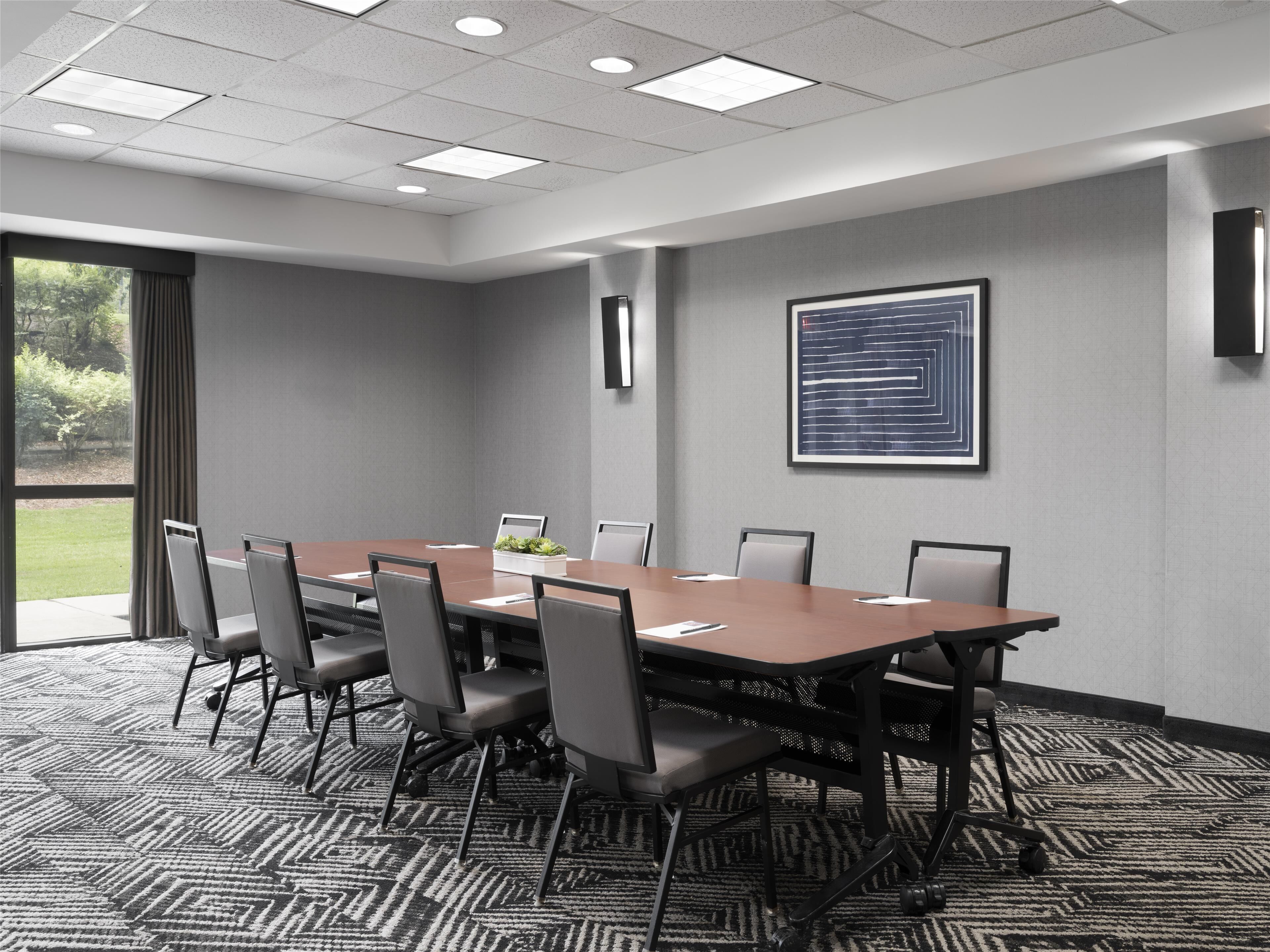 A conference room with a long table, chairs, a potted plant, and a framed artwork on the wall.