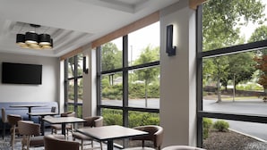 Un espace repas moderne, doté de tables et de chaises, de grandes fenêtres offrant une vue sur l’extérieur et d’un téléviseur à écran plat fixé au mur.