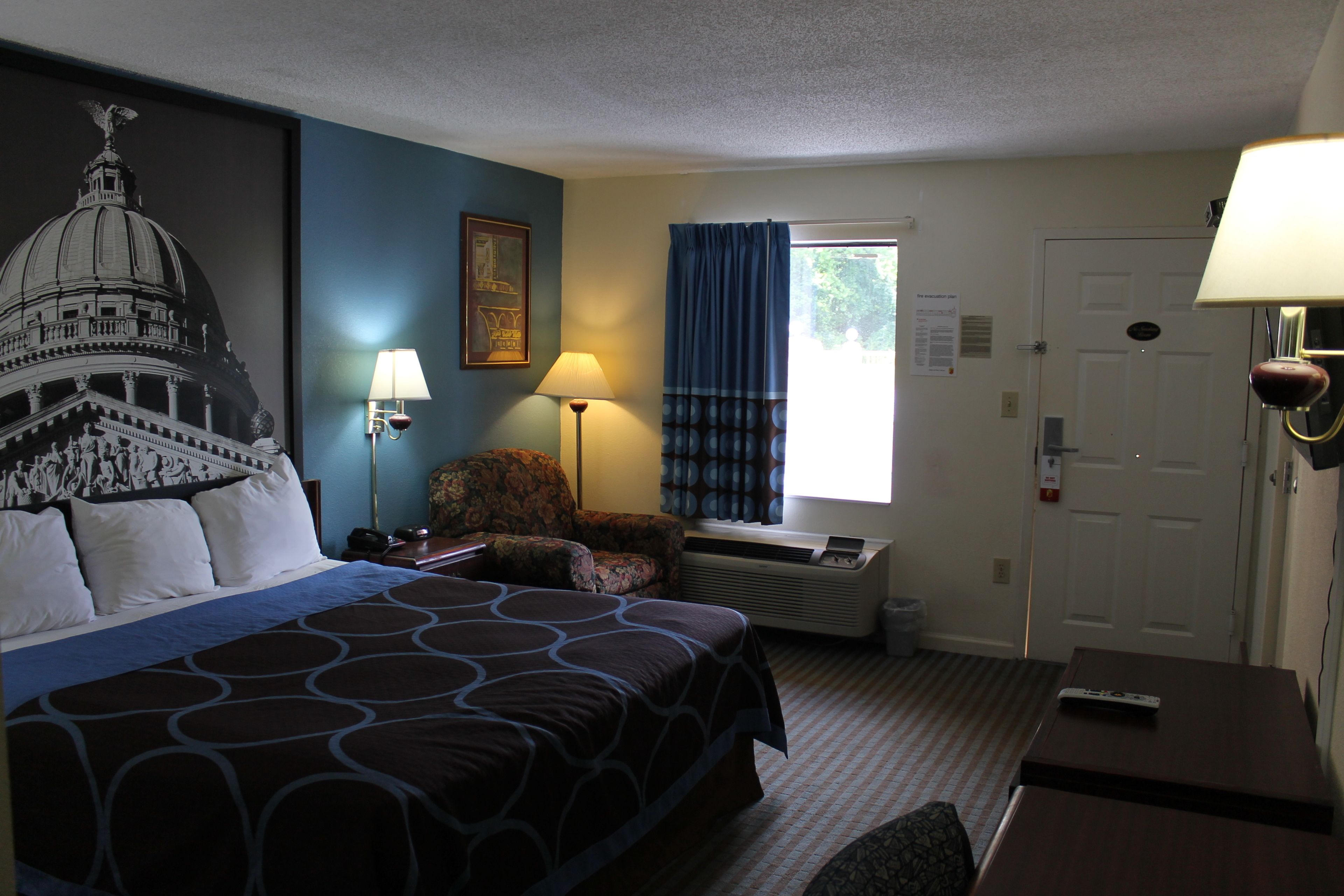 A hotel room with a large bed, a desk, a chair, a lamp, a window with curtains, and a wall-mounted air conditioner.