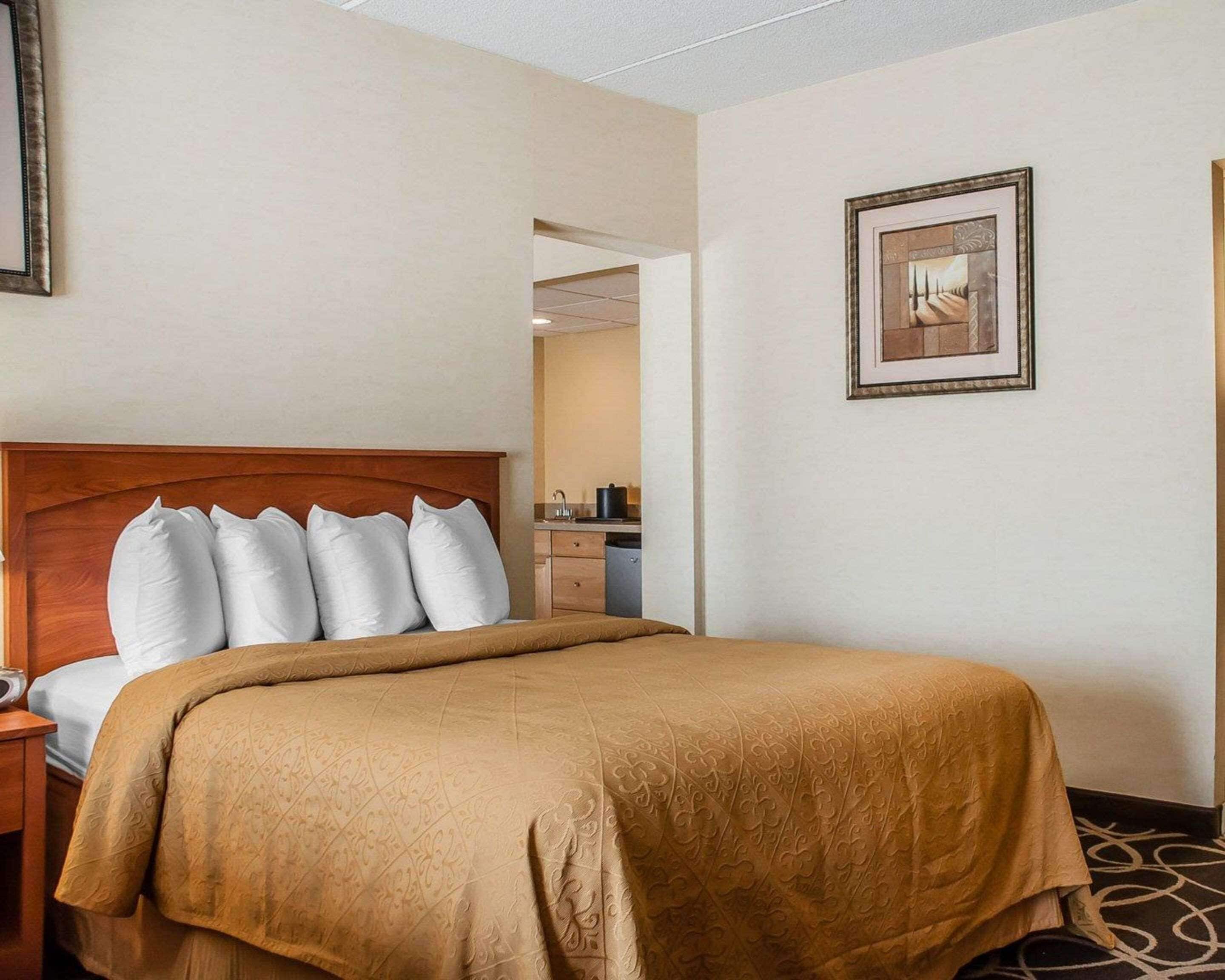 A hotel room with a bed, a nightstand, a framed picture on the wall, and a view into the adjacent room.