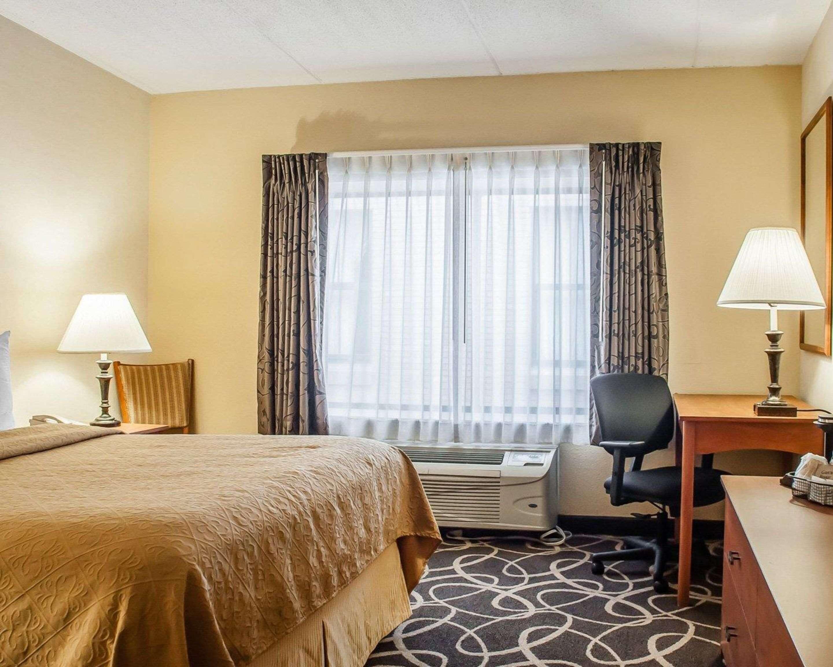Una habitación de hotel con cama, escritorio, silla, equipo de aire acondicionado y ventana con cortinas.