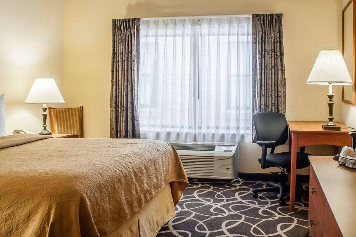 Una habitación de hotel con cama, escritorio, silla, equipo de aire acondicionado y ventana con cortinas.