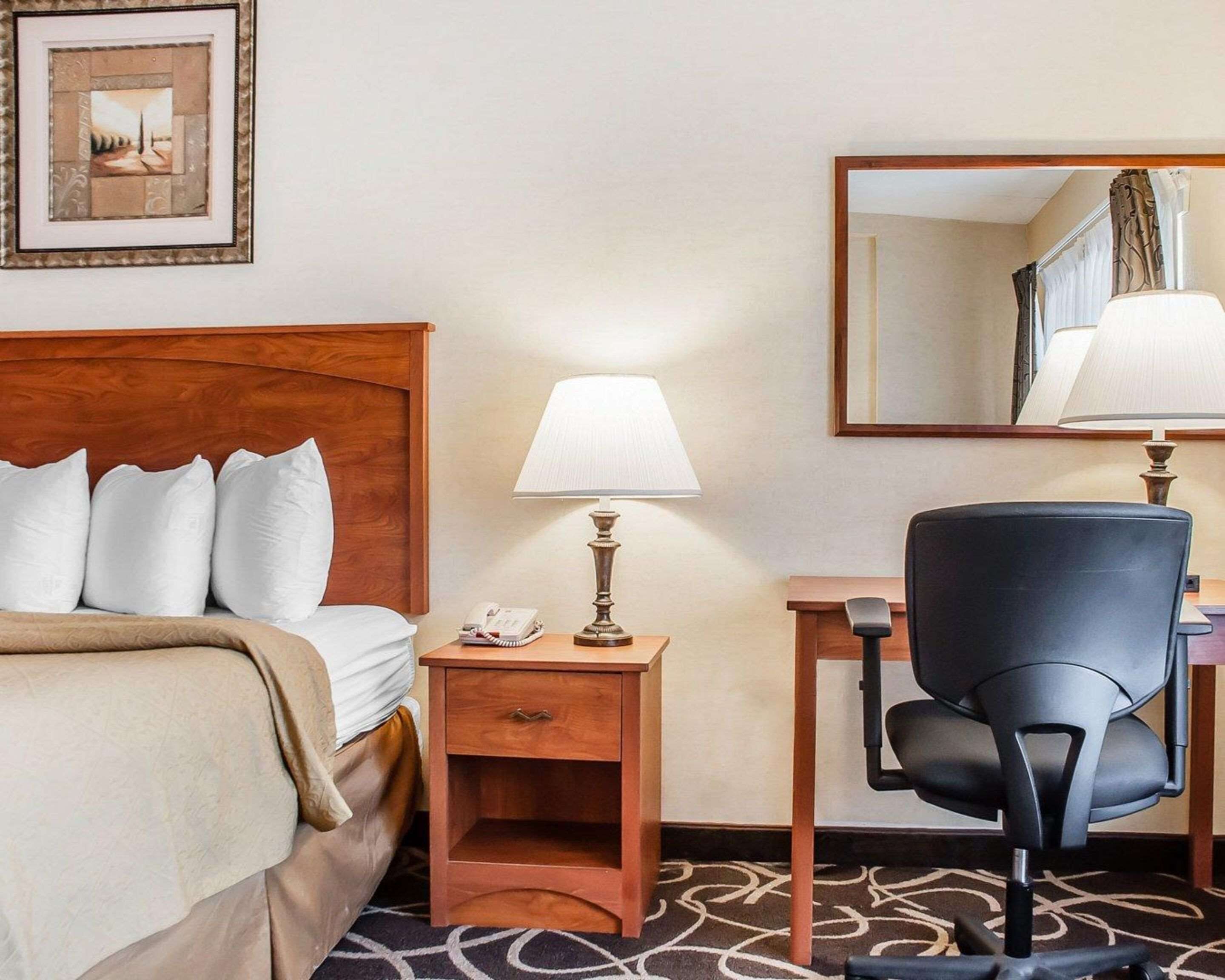 A hotel room with a bed, a desk, a chair, a nightstand with a lamp, a telephone, and a framed picture on the wall.