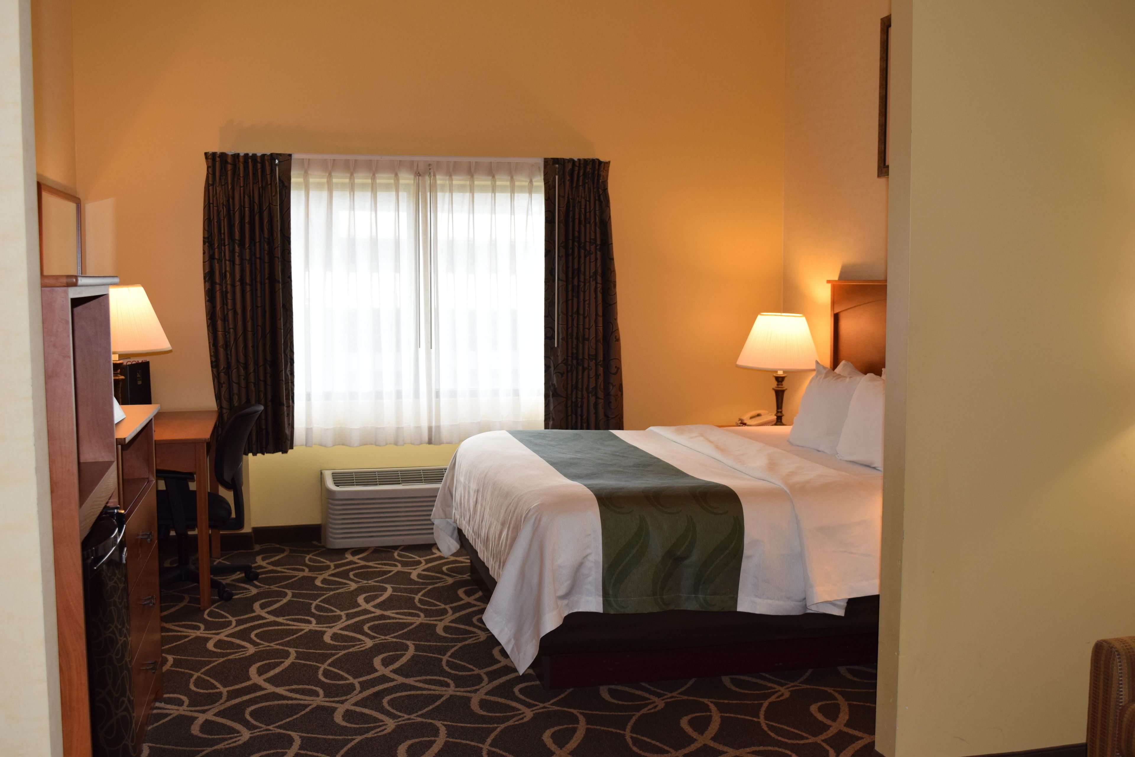 A hotel room with a bed, two bedside lamps, a desk with a chair, and a window with curtains.