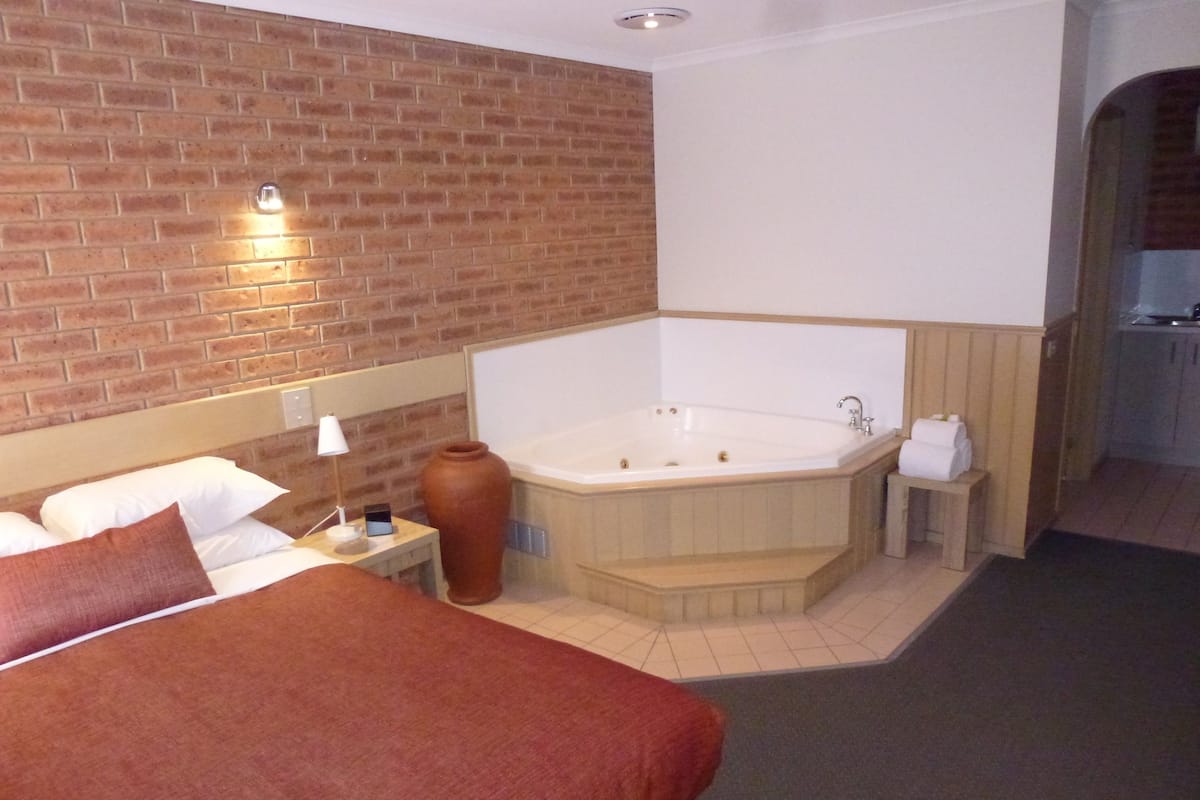 A hotel room with a brick wall, a large bed with a red blanket, a white bathtub, and a small bedside table with a lamp and a vase.