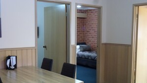 Quarto com mesa e cadeiras de madeira, cama e parede de tijolos ao fundo, e porta que dá para outro cômodo.
