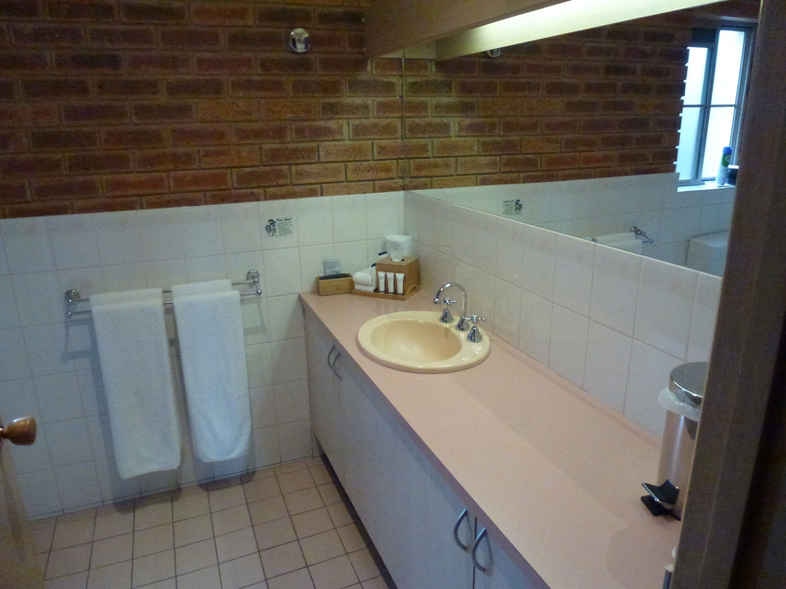 A bathroom with a beige sink, white tiles, a mirror, and a brick wall.