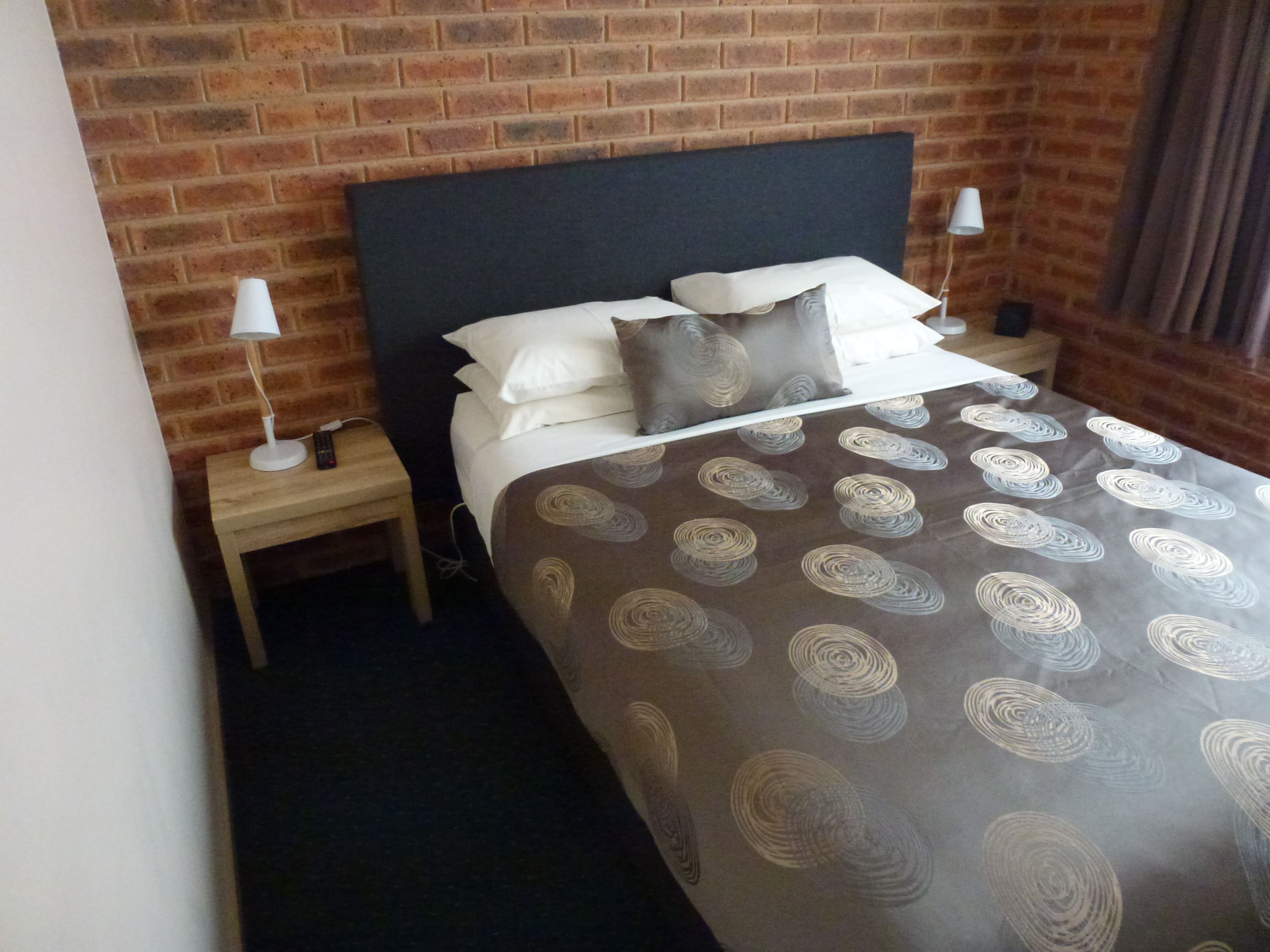 Une chambre d’hôtel avec un mur en briques, un grand lit recouvert d’une courtepointe à motifs, deux tables de chevet avec des lampes et un rideau à la fenêtre.