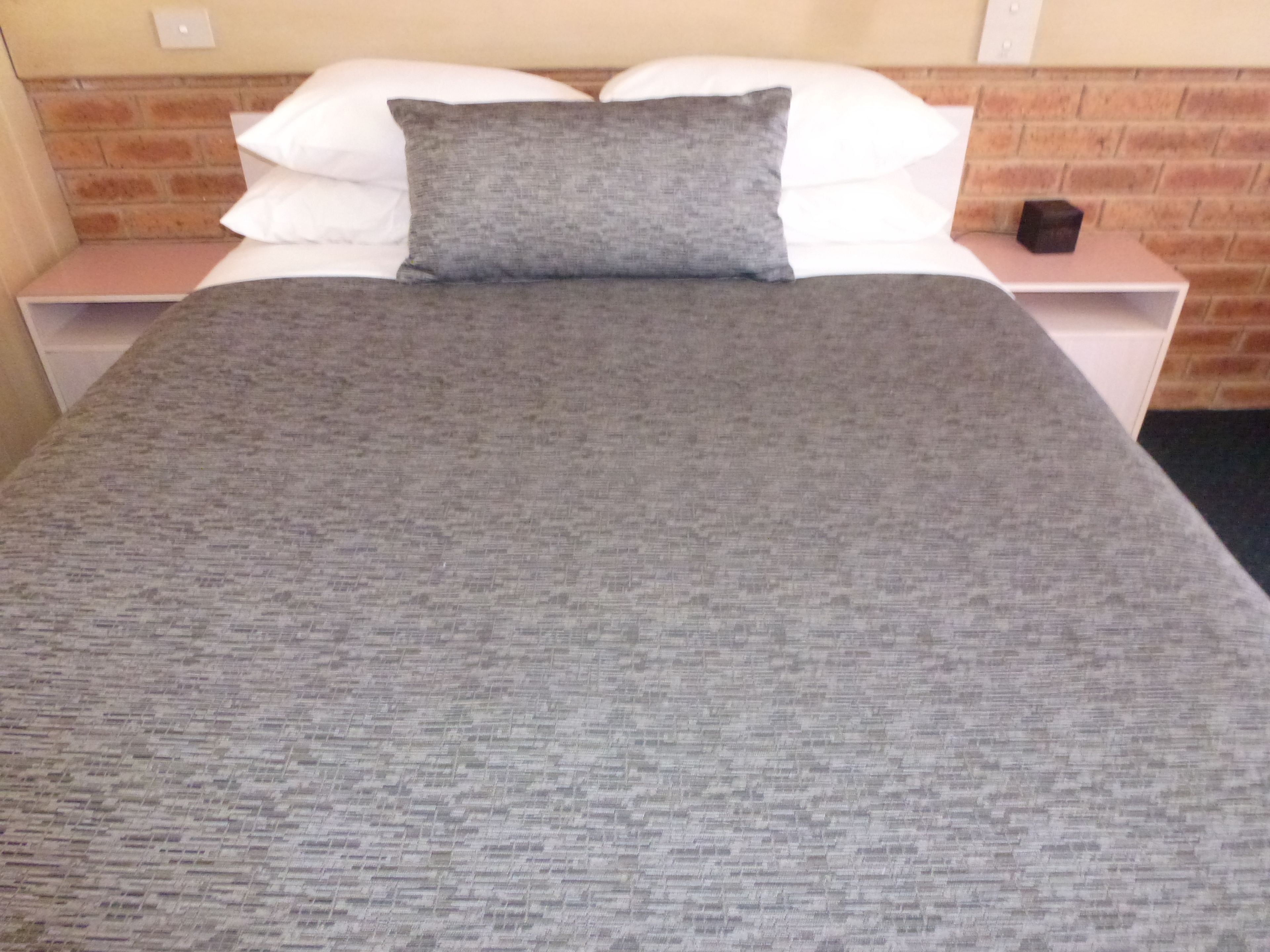 A neatly made bed with a grey blanket and pillows, a brick wall behind, and a small bedside table with a lamp.