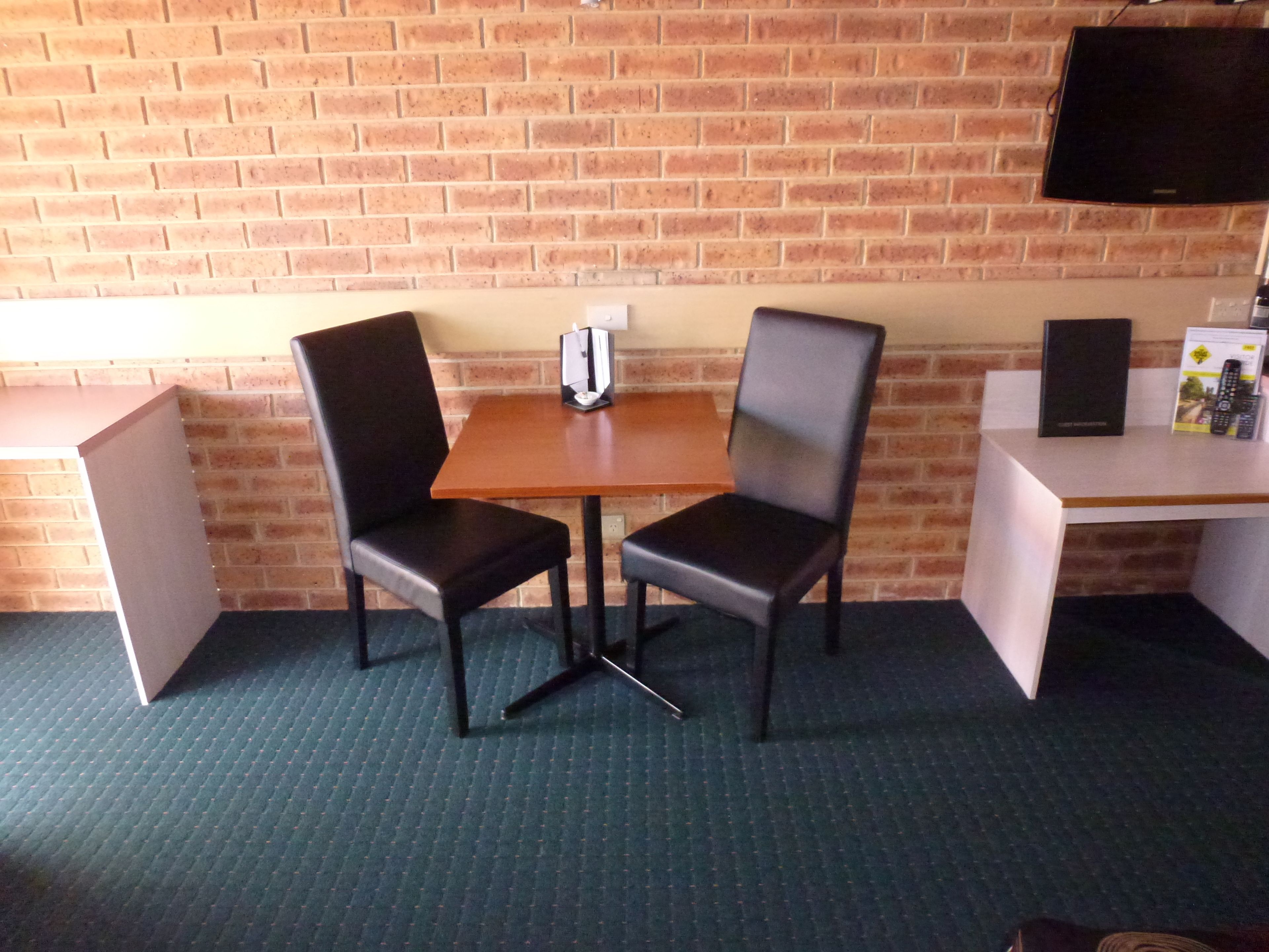 A room with a brick wall, a wooden table with two black chairs, a whiteboard, a television, and a small desk with a speaker and remote controls.