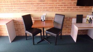 A room with a brick wall, a wooden table with two black chairs, a whiteboard, a television, and a small desk with a speaker and remote controls.