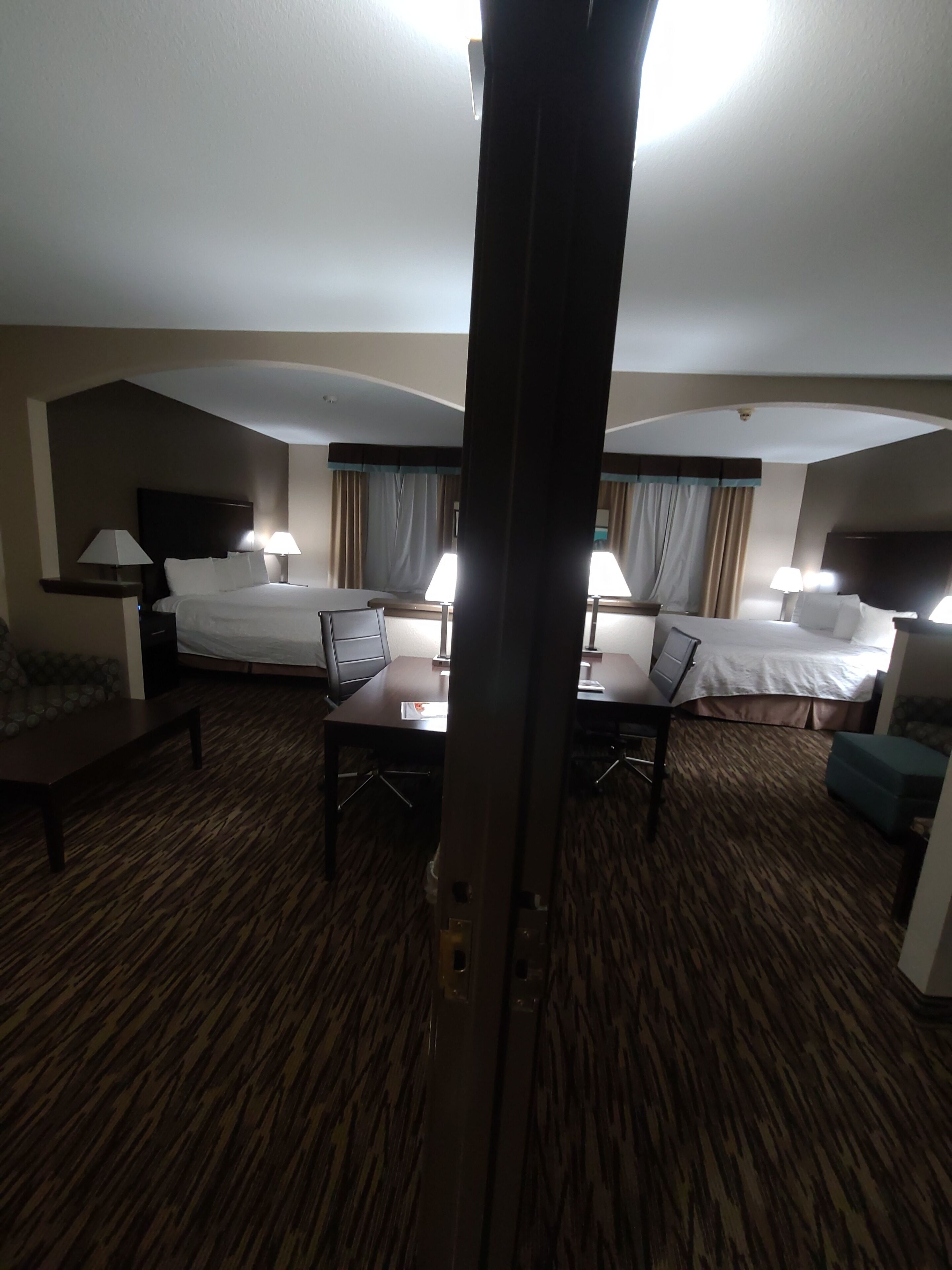 A hotel room with two beds, a desk, and a chair. There are lamps on either side of the beds and a window with curtains.