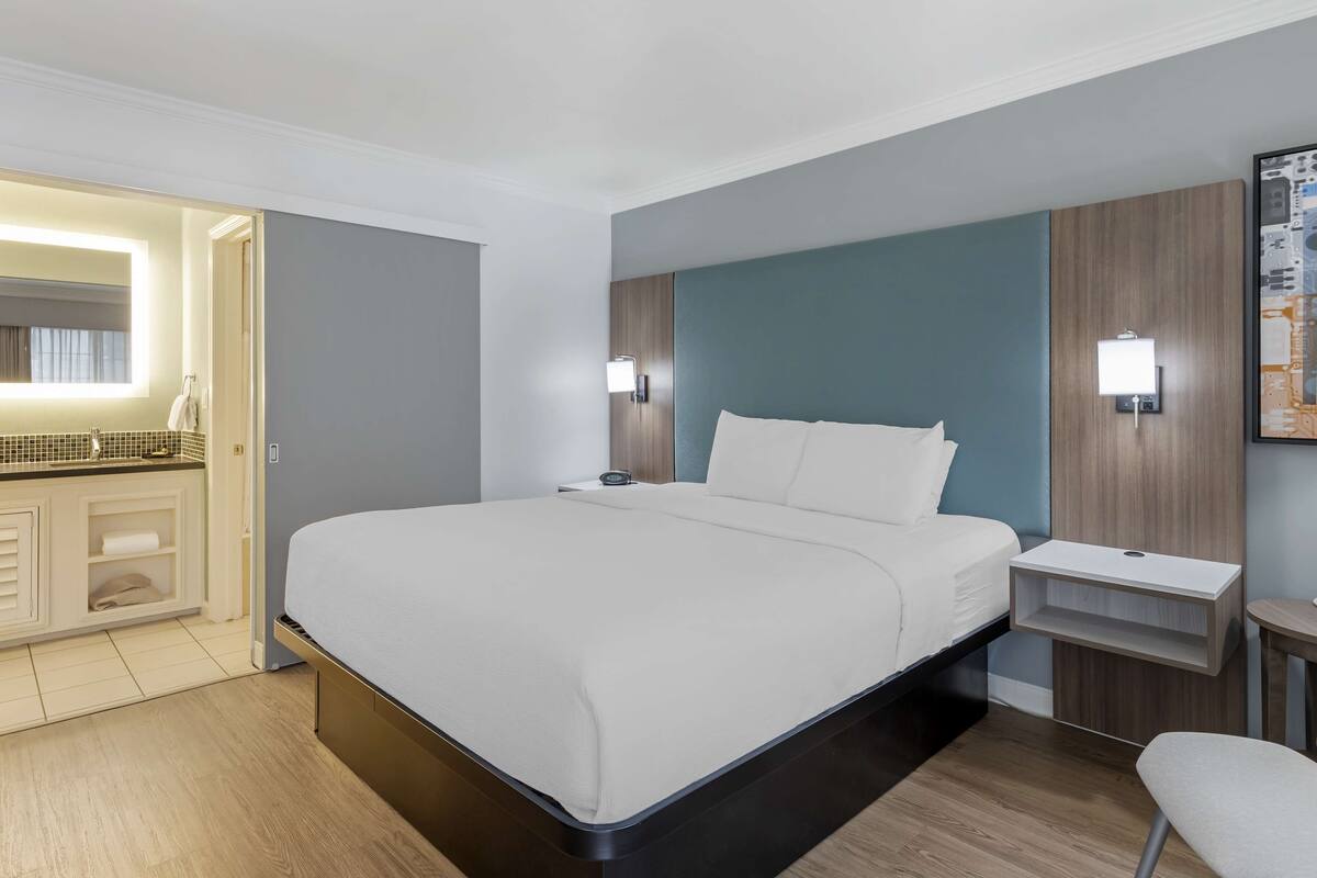A modern hotel room with a large bed, bedside tables, a small desk, and a bathroom visible through an open door.