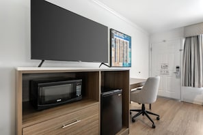 A hotel room with a flat-screen TV mounted on the wall, a microwave, a small refrigerator, a grey office chair, and a wooden desk.