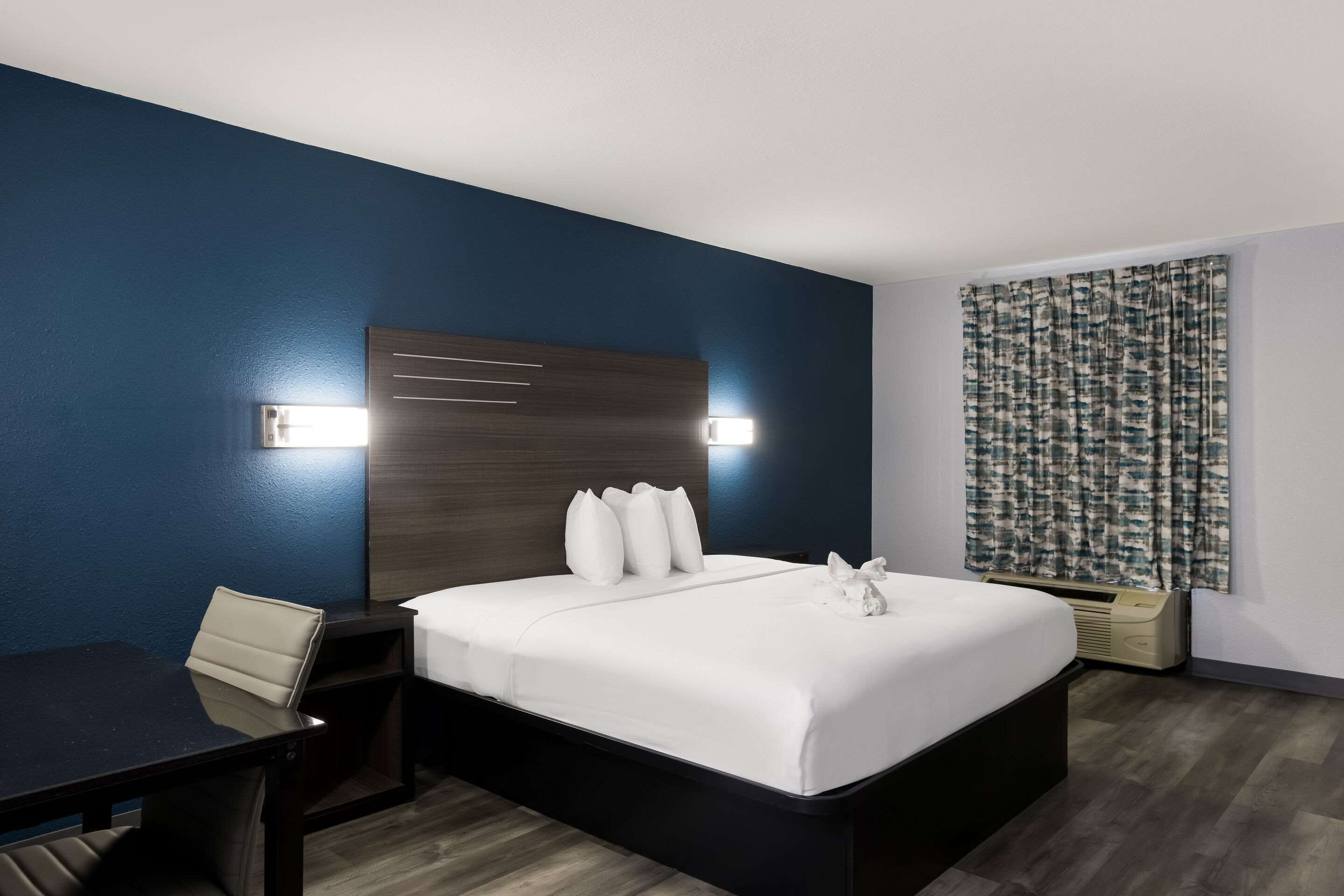 Una habitación de hotel moderna con una pared de acento azul oscuro, un cabecero de madera, una cama blanca con almohadas, una mesita de noche y un escritorio.