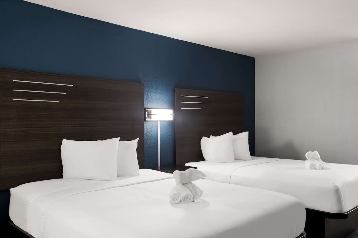 A hotel room with two beds, dark wood headboards, white bedding, and a bedside lamp.