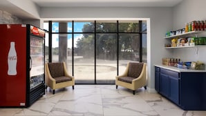 A room with a Coca-Cola refrigerator, two patterned armchairs, a large window, and a shelf with snacks. - Suburban Studios Hinesville - Fort Stewart Area (Hinesville)